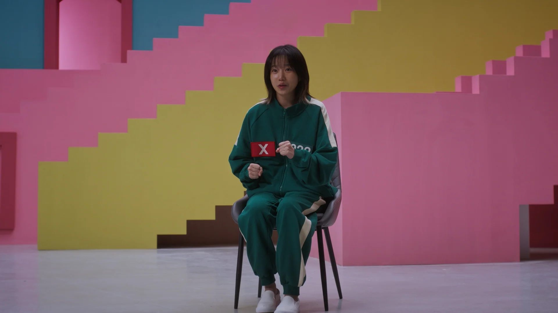 Inside Squid Game S2 — a player actress seated wide on the pastel staircase set