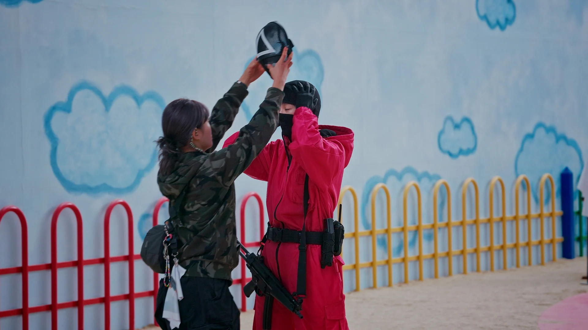 Inside Squid Game S2 — crew fitting a guard mask beside the painted fence